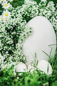 weiße Eier vor großem Ei neben weißen Blüten im Gras