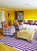 Rustic wooden coffee table, tartan armchairs and purple and white striped rug in living room with yellow walls