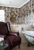 Free-standing bathtub, leather armchair, mirror and wallpaper with pattern of palm trees in bathroom