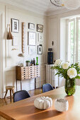 View past flowers on dining table to chest of drawers below gallery of pictures on wall