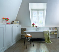 Free-standing bathtub, stool and fitted cupboards below shelf in white bathroom with dormer window