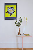 Vase of leaves and reading lamp on side table below colourful artwork