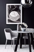 Vase with orchid and glass bowl on round table with marble top and armchair against black wall with picture