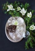 Glass Christmas-tree baubles and pine cones on silver platter next to green conifer branches and hellebore flowers