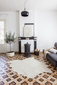 Cowhide rug on patterned tiled floor, grey sofa, wood-burning stove in front of disused fireplace and potted olive tree in living room