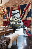 Sheepskins on bench at dining table, brick wall and Union-flag-patterned curtains screening open doorway