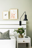 Vase of flowers on white bedside table next to bed with white-painted wooden headboard