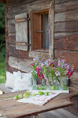 Phlox, love-in-a-mist and snapdragons in small bottles decorating table outside rustic wooden farmhouse