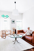 Blonde Frau auf cognacfarbener Ledercouch, Couchtisch mit Glasplatte und Designer-Rattanstuhl im Wohnzimmer mit weißen Wänden