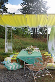 Set table with green tablecloth, chair, tree-stump stools and cushions under awning
