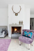 Scatter cushion on antique easy chair with lilac upholstery next to fireplace below antlers on wall
