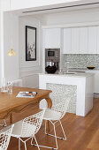 Wooden table and white classic chairs, island counter and white fitted kitchen cabinet in open-plan interior
