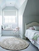 White bed under sloping ceiling, round carpet and built-in bench under window in girl's room