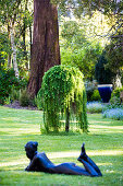 Liegende Frauenskulptur auf Rasenfläche, im Hintergrund japanische Hängelärche (Larix decidua 'Pendula') im Garten