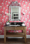 Washstand with marble top below silver-framed mirror on wall with flamingo-patterned wallpaper