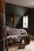 Double bed in rustic attic room with wooden columns, tiled stove and dark walls