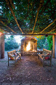 Round table and wooden benches below climber-covered pergola in front of open fire in fireplace