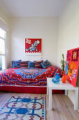 Colourful bedspread and scatter cushions on bed below comic-style artwork behind vases and felt collage on side table