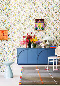 Blue sideboard with bouquet and table lamp in front of floral wallpaper