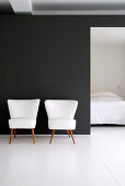 Zwei weiße Retro-Sessel vor schwarzer Wand, Blick ins Schlafzimmer