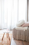 Fringed blanket and cushions on ottoman and stool in front of window with floor-length curtains
