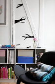 Black chair with cushions in front of a shelf with books, above a stylized branch as a wall decoration