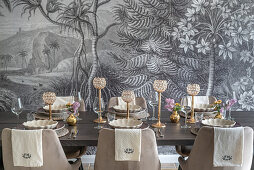 Festively set dining table with upholstered chairs in front of monochrome mural wallpaper
