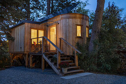 Wooden tiny house on stilts with exterior steps and small veranda at twilight illuminated from within