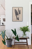 A house plant next to a small table with a flower and a picture hanging above it