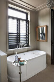 White, free-standing bathtub below window with louvre blinds in bathroom