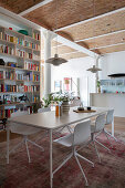 White dining table with chairs, above pendant lamp in front of wall of books in open living room with high ceiling
