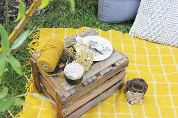 Wooden box with spool of yarn, jam jar, flower, plate and napkin on yellow picnic blanket