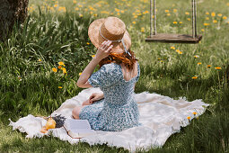 Female in dress and straw hat sitting on picnic blanket on green meadow near swings in summer countryside