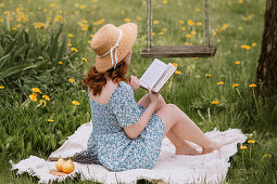 Female in dress and straw hat reading novel while sitting on picnic blanket on green meadow near swings in summer countryside