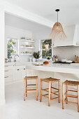 White kitchen with wooden stools around kitchen island, rattan pendant lights, open shelves in the background