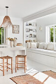 White kitchen with wooden stools around kitchen island, rattan pendant lights and built-in bench in front of window