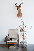 Armchair next to vase of dried grasses under antelope head on wall; autumnal ambience
