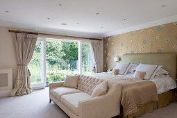A double bed with a high headboard in front of a wallpapered wall and a light sofa in a bedroom