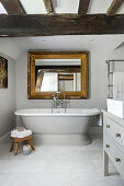 Frei stehende Badewanne und Goldrahmenspiegel in hellgrauem Bad mit rustikalen Holzbalken