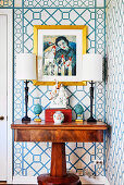 Antique table with lamps, above it wallpaper with geometric pattern and painting