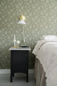Bedside table and wall lamp next to bed in bedroom with floral wallpaper