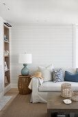 Helles Sofa mit Kissen und Beistelltisch mit Lampe vor weiß gestrichener Holzwand