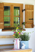 Frühlingsstrauß aus Fresien und Tulpen, daneben Topf mit Rosmarin und Osterhase auf Bank vor Sprossenfenster