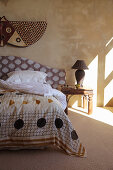 Double bed with geometric patterned headboard and antique bedside table in a sunlit bedroom