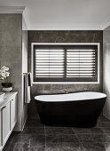 Badezimmer in dunklen Tönen mit schwarzen Marmorfliesen, freistehender, schwarzer Badewanne und Fensterläden