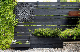 Schwarz gestrichener Holzzaun mit dekorativem Element, kleinem Wasserbecken und Pflanzkasten