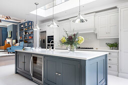Weiße Einbauküche, davor Kücheninsel mit blau-grauen Fronten und weißer Arbeitsplatte in offenem Wohnraum
