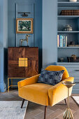 Elegant highboard and yellow armchair in front of blue wall next to shelving unit