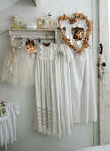 Weiße Kleidungsstücke mit romantischen, femininen Details am Wandregal, alte Weihnachtskarten und herzförmige Kranz mit getrockneten Blüten