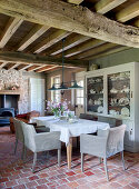 Esstisch und Stühle mit Leinenbezug vor Geschirrschrank im Zimmer mit Terrakottafliesenboden und Holzbalkendecke